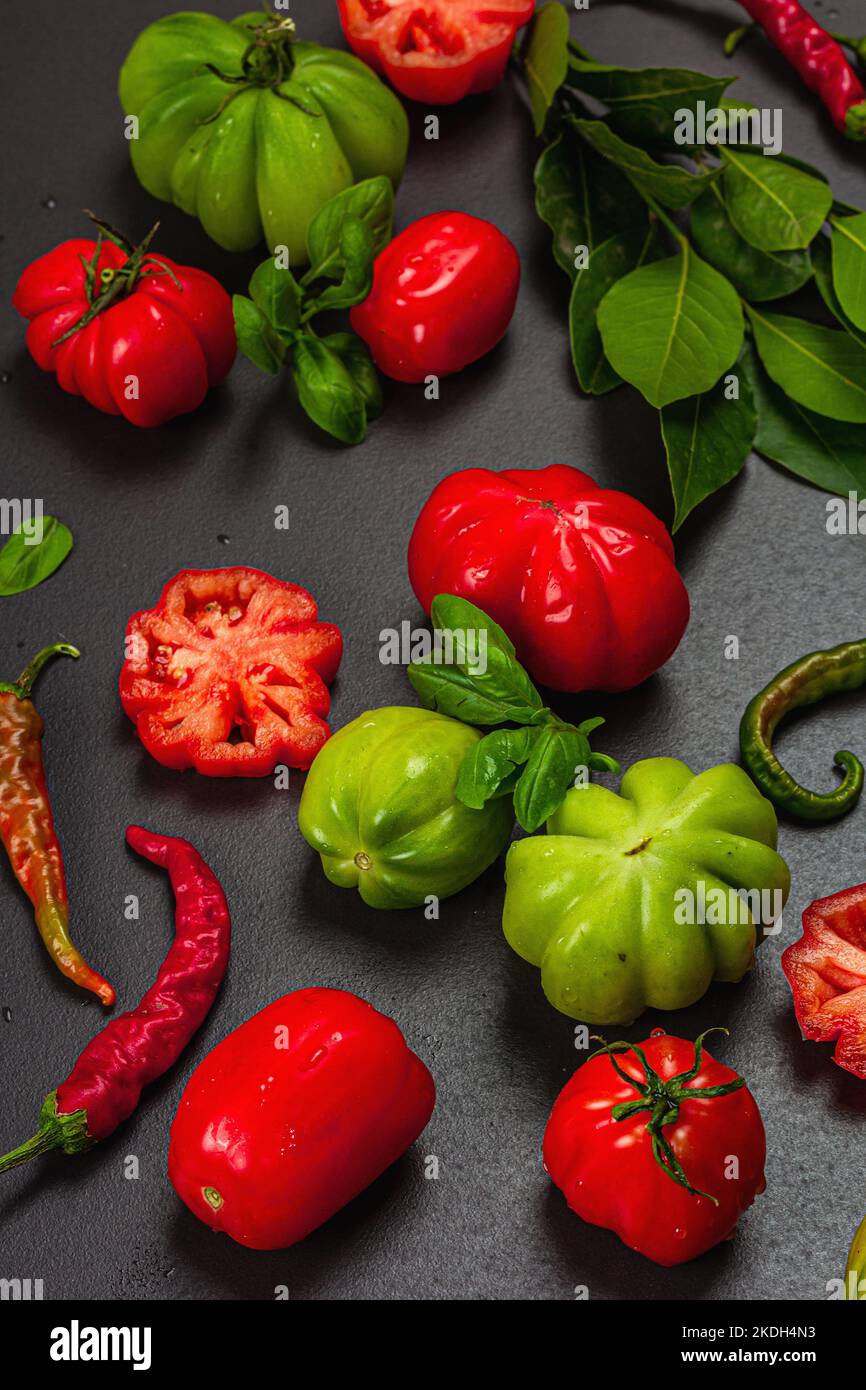 Pomodori verdi e rossi a coste, varietà americana o fiorentina. Verdure mature, peperoncino, foglie fresche di alloro. Luce tenebrosa, ombra scura, calcestruzzo di pietra Foto Stock