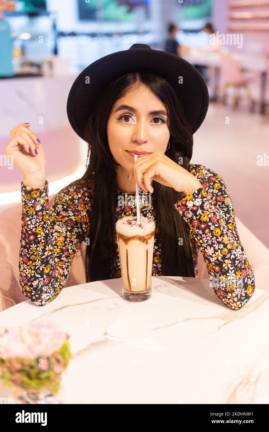 giovane donna in un cappello che tiene una paglia da un bicchiere con frullato, bevanda rinfrescante nel ristorante, lifestyle che si diverte Foto Stock