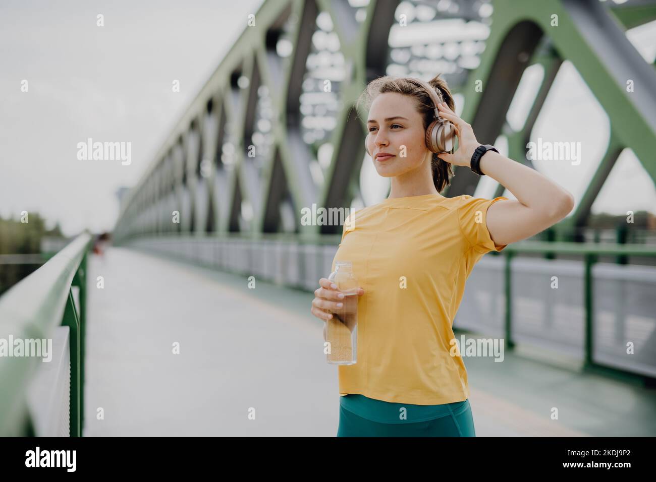 Giovane donna che beve acqua e ascolta musica con le cuffie, durante il jogging in città, stile di vita sano e concetto sportivo. Foto Stock