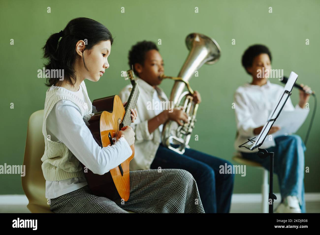 Simpatica studentessa asiatica che suona la chitarra acustica e guarda le note in piedi mentre siede contro due compagni di classe a lezione Foto Stock