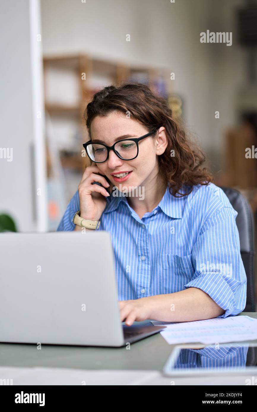 Giovane donna che parla al telefono utilizzando un computer portatile che lavora online. Verticale Foto Stock