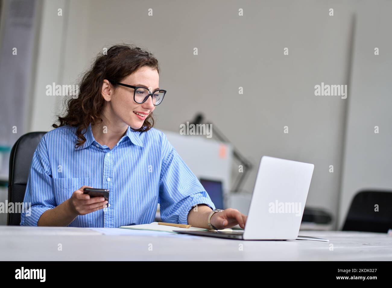 Giovane donna d'affari seduta alla scrivania che lavora online su un laptop che tiene il telefono. Foto Stock