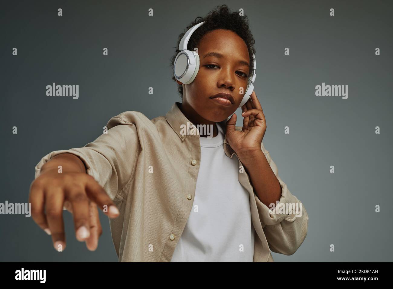 Carino ragazzo afro-americano in cuffie guardando la fotocamera mentre si è in piedi in isolamento su sfondo grigio e godersi la musica rap Foto Stock