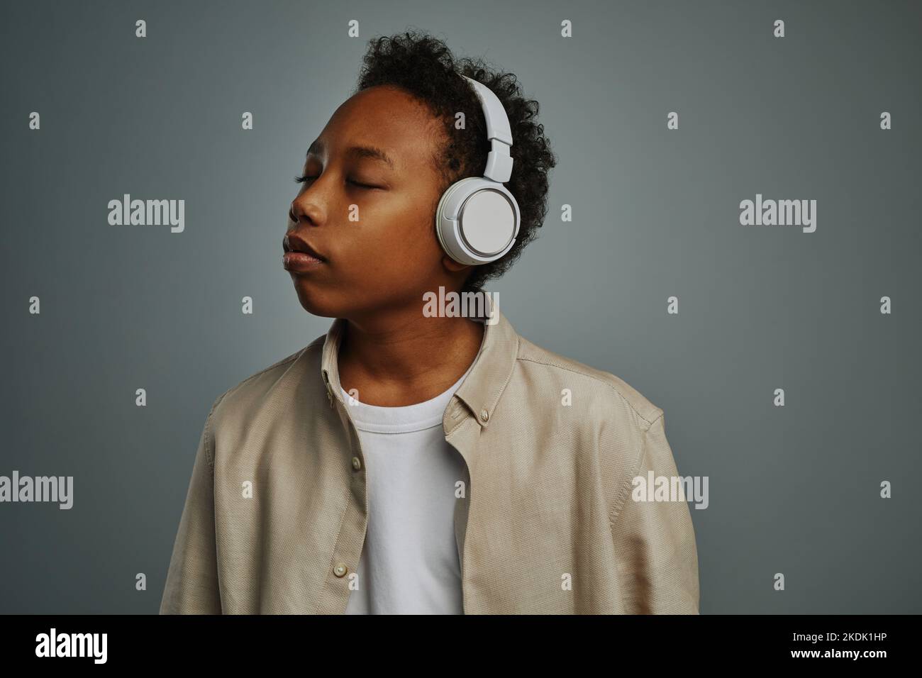 Adorabile ragazzo sereno che ascolta la musica rilassante con cuffie bianche mentre tiene gli occhi chiusi e si posa per la fotocamera su sfondo grigio Foto Stock
