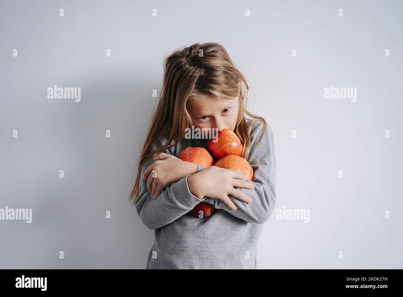 Ragazzo pio che abbraccia un mazzo di mele tra le braccia. Su una parete bianca. Ha lunghi capelli biondi. Foto Stock