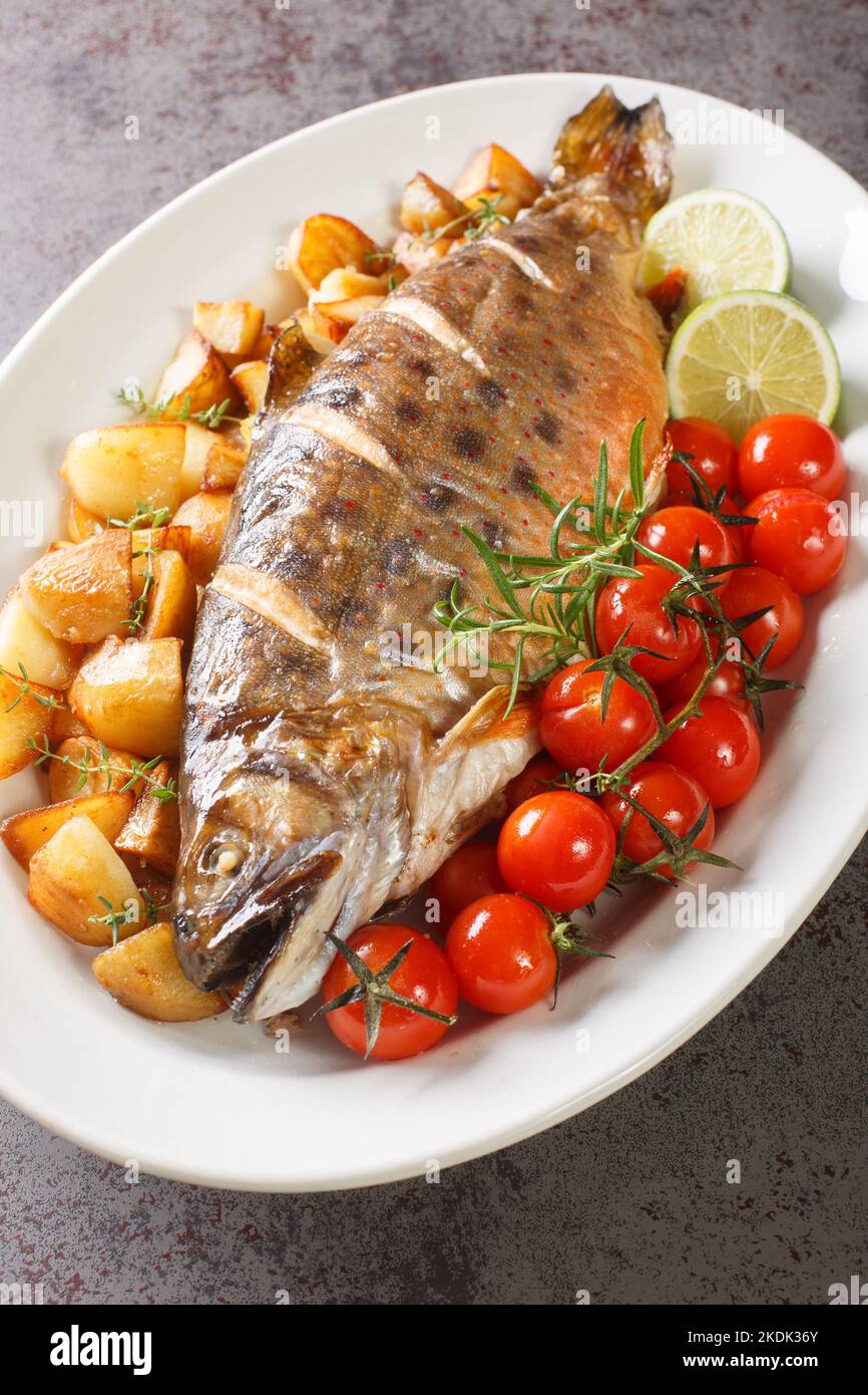Pesce intero cotto al forno con patate fritte, pomodori ciliegini e lime primo piano su un piatto sul tavolo. Verticale Foto Stock