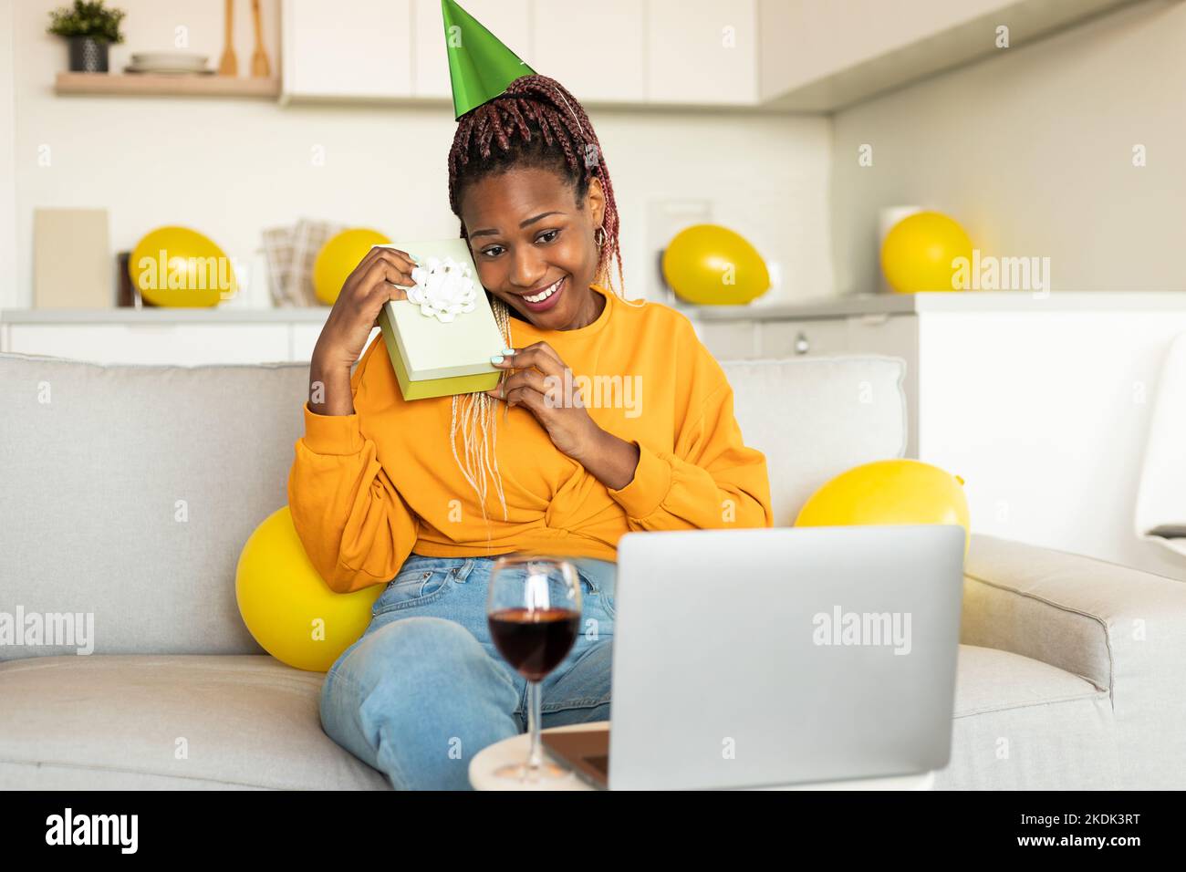 Donna afro-americana positiva che tiene presente mentre ha b-day party online a casa, guardando il notebook e sorridendo Foto Stock