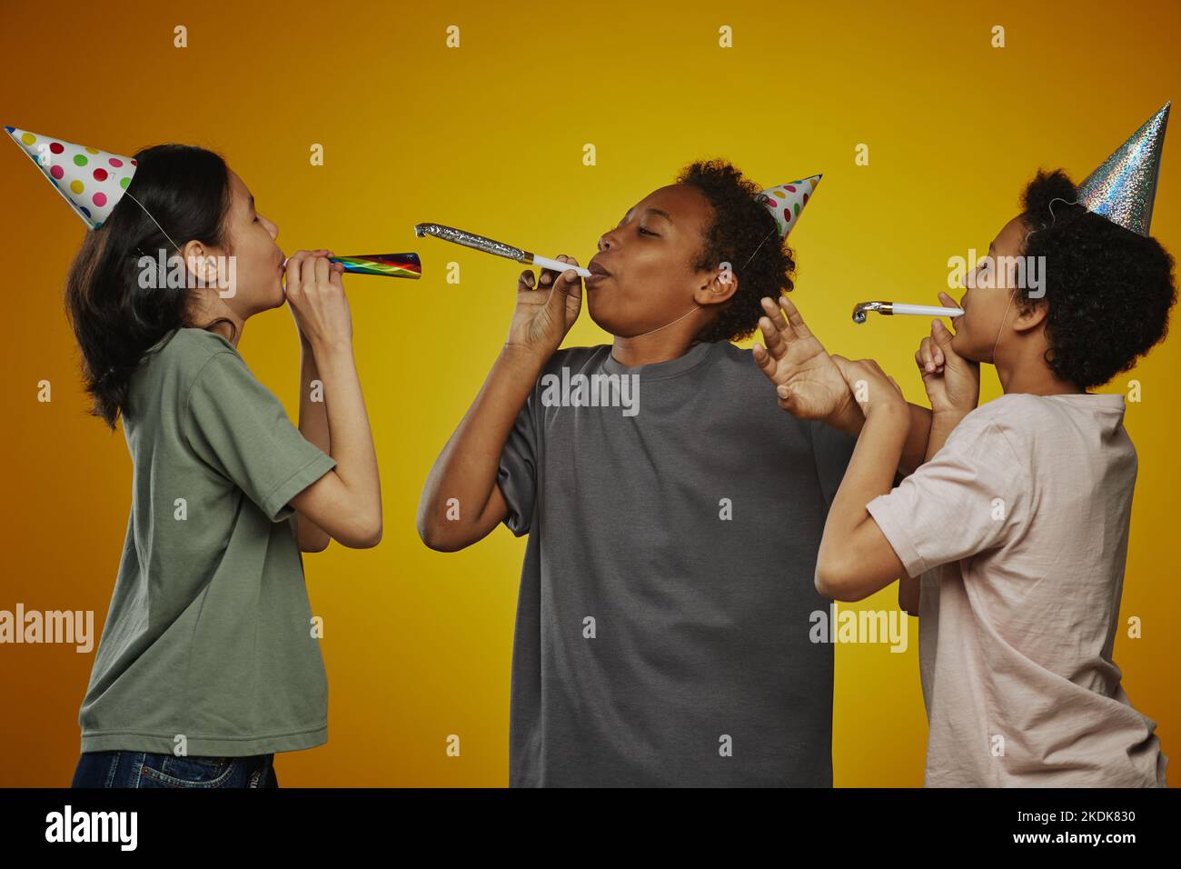 Gruppo di tre bambini pre-teen interculturali in t-shirt e cappellini di compleanno che soffiano fischietti mentre si diverte e si diverte Foto Stock