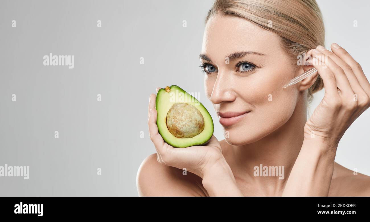 Donna caucasica che applica olio di avocado sul viso con contagocce per la pelle nutriente e idratante e tiene metà di avocado, di alta qualità. Fa Foto Stock