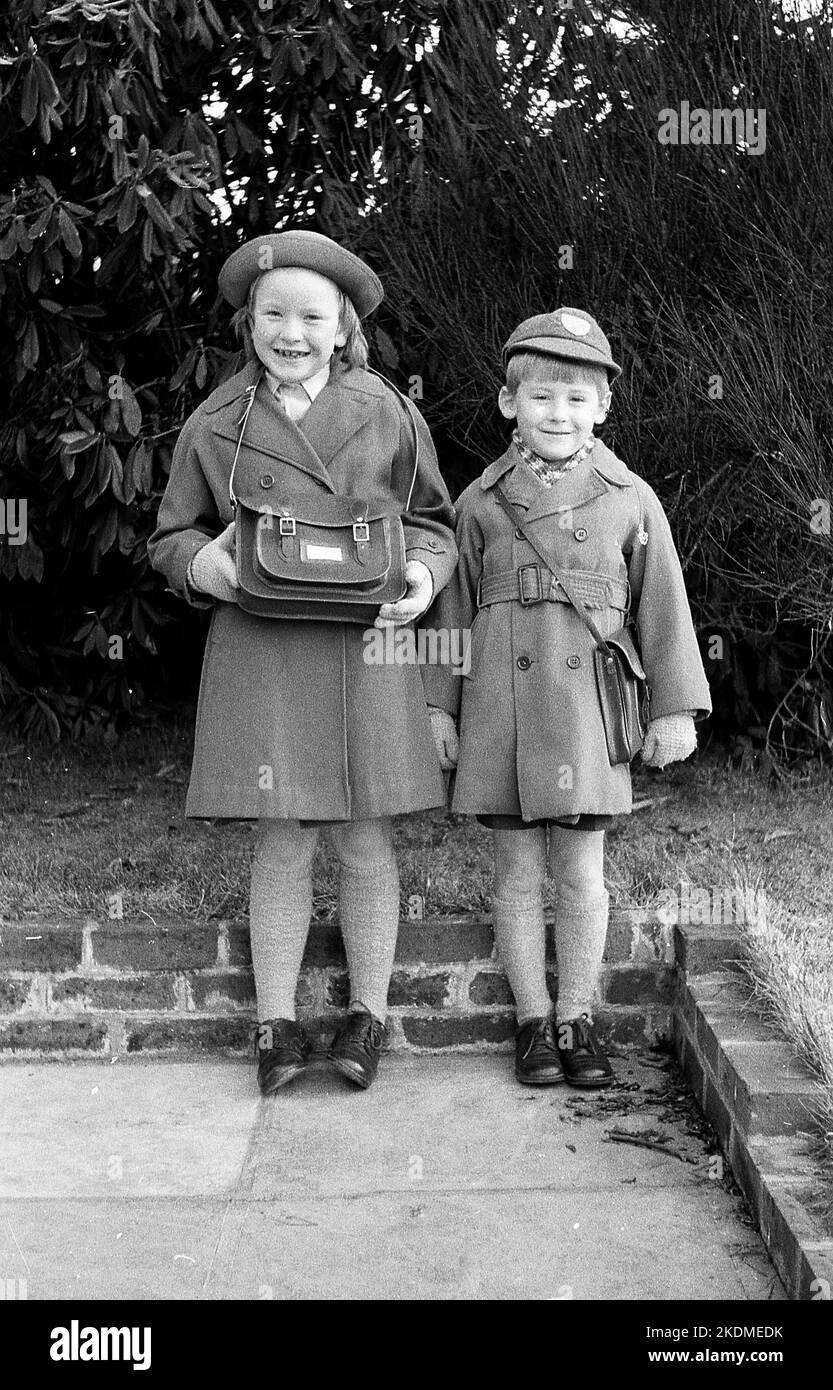 1963, storico, fratello e sorella stanno insieme per una foto fuori nella loro divisa scuola, cappotti e cappelli, la ragazza orgogliosamente tenendo la sua borsa a mano scuola, Inghilterra, Regno Unito, Foto Stock