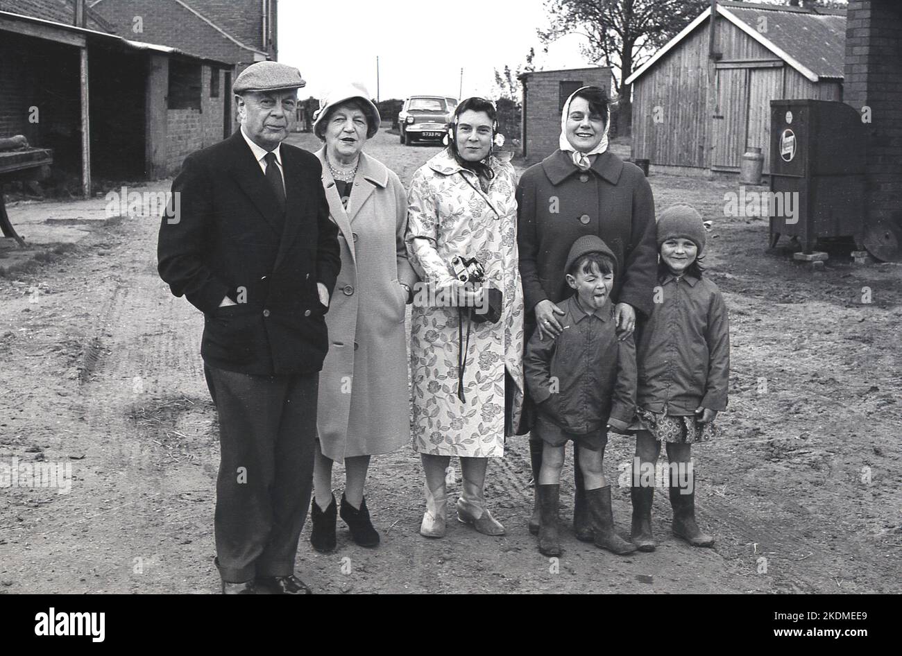 1960s, storico, in piedi insieme per una foto in un cortile, una famiglia, due nonni, una madre più la sua amica e due bambini piccoli, in una visita ad una fattoria, il ragazzo non impressionato..Tongue out,,,YUK! Inghilterra, Regno Unito. Foto Stock
