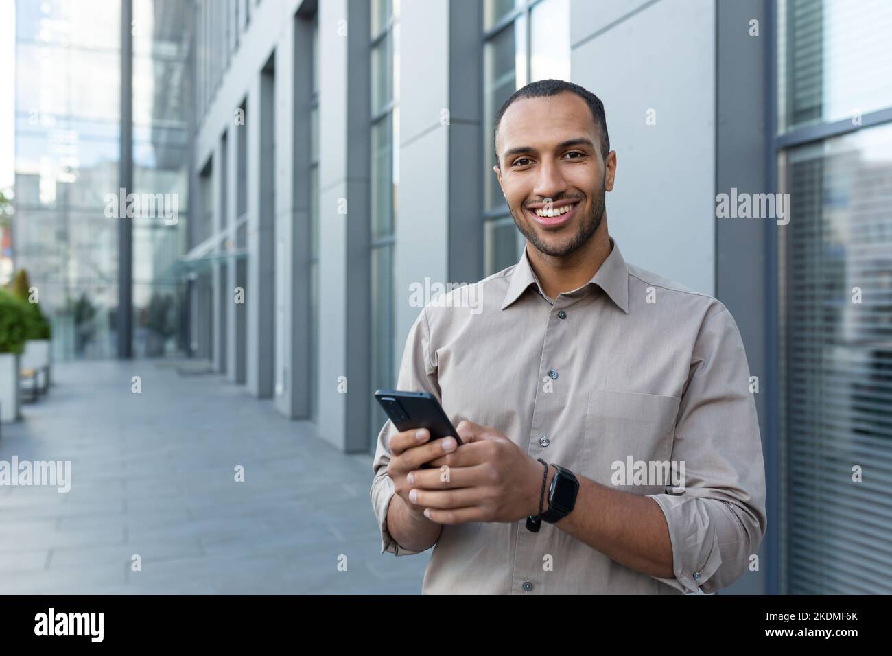 Ritratto dell'uomo d'affari afro-americano, uomo che sorride il telefono e guarda la macchina fotografica, impiegato in camicia fuori dal moderno edificio d'ufficio durante la pausa pranzo. Foto Stock