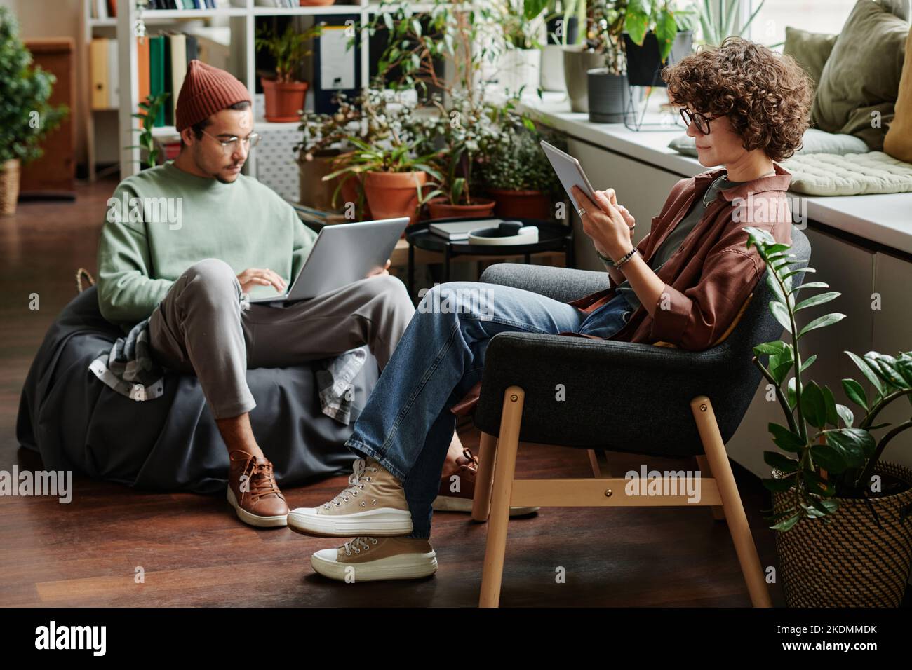 Due giovani colleghi seri che utilizzano gadget mobili senza fili mentre si siedono in poltrone in ufficio open space con varietà e piante verdi Foto Stock