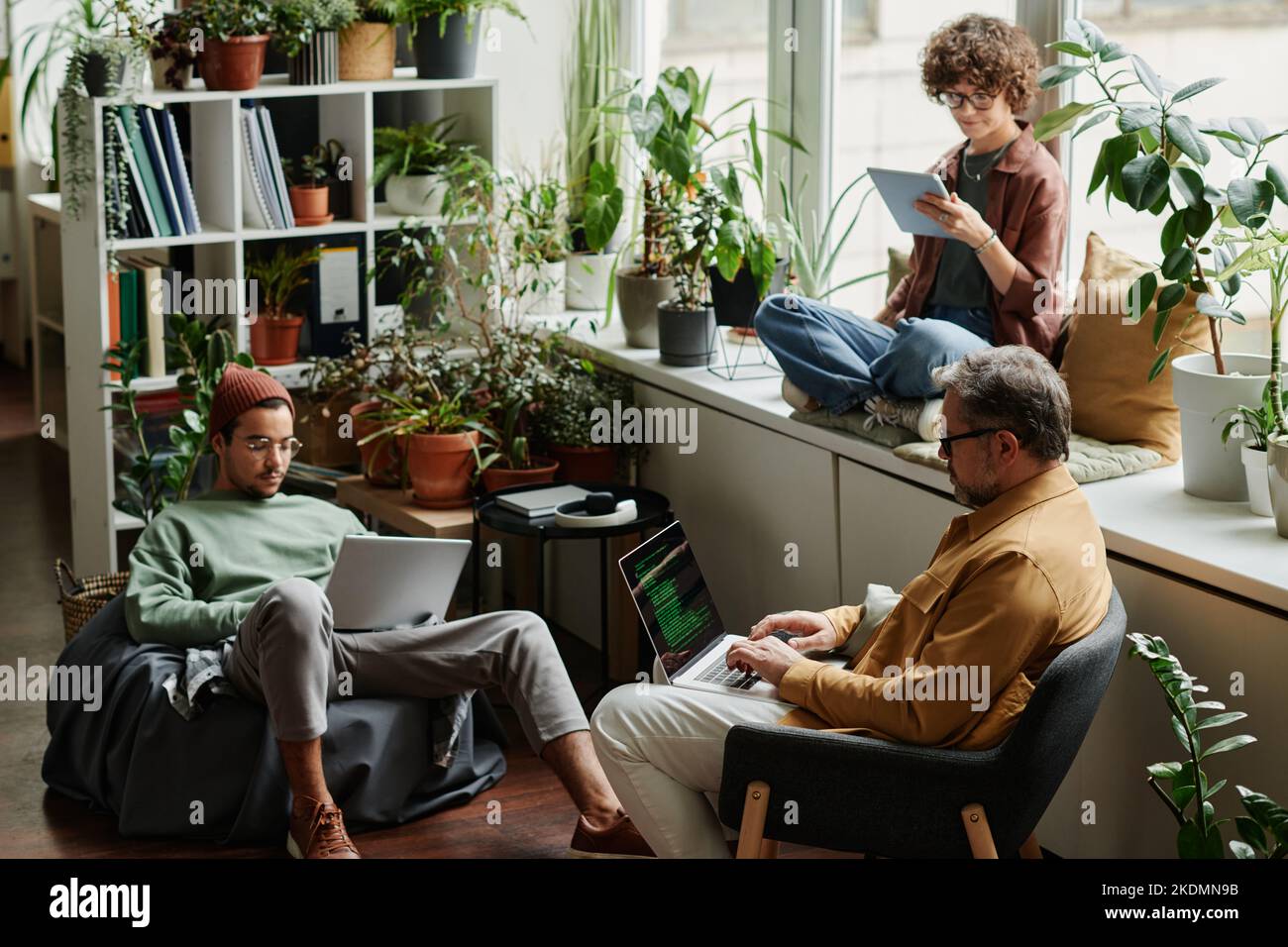 Tre colleghi multiculturali in abbigliamento casualwear che utilizzano gadget mobili in ufficio mentre un tecnico IT maturo decodifica i dati sullo schermo del notebook Foto Stock