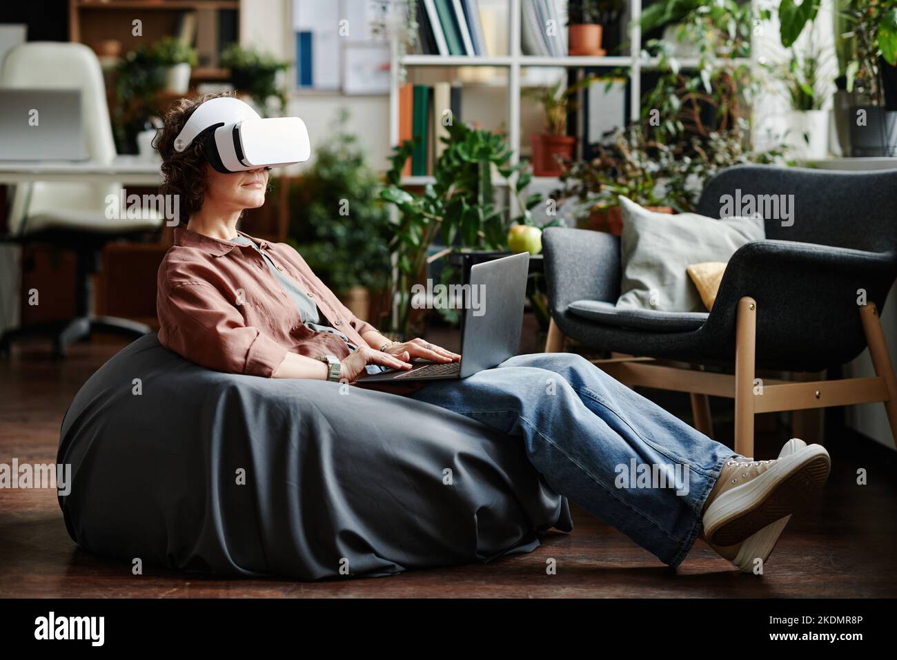 Giovane donna d'affari moderna con visore vr e computer portatile in ginocchio seduto sulla poltrona e in preparazione alla presentazione del nuovo progetto Foto Stock
