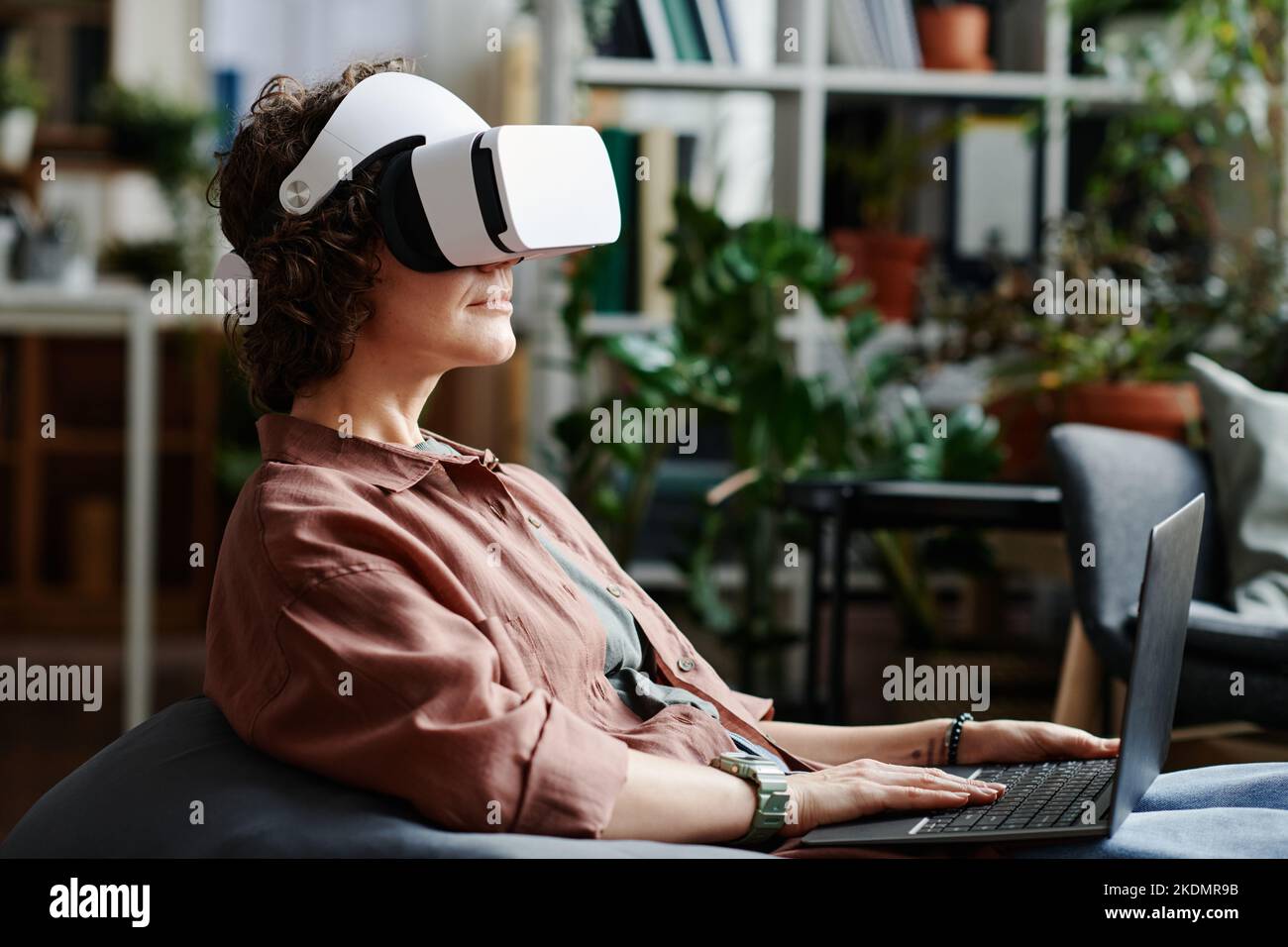 Vista laterale di una giovane programmatore in visore vr che utilizza un computer portatile per controllare il lavoro con oggetti virtuali mentre si è seduti in un ufficio open space Foto Stock