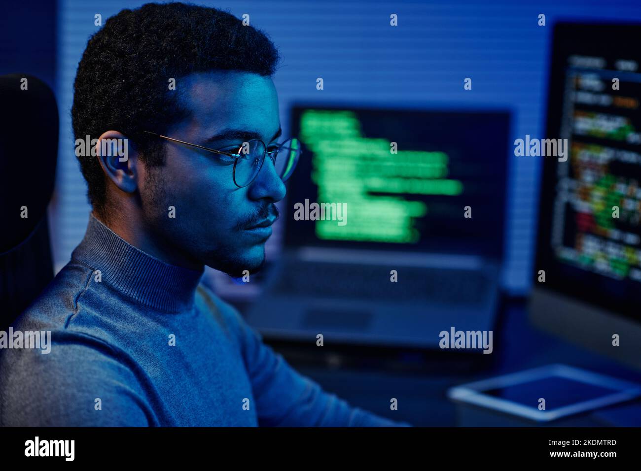 Tecnico di supporto IT giovane e serio negli occhiali che guardano lo schermo del computer mentre si siede davanti ai monitor e decodifica i dati Foto Stock