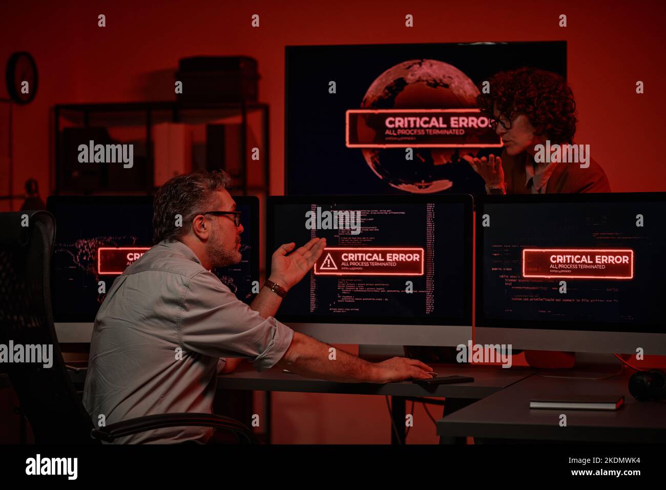 Programmatore maturo seduto di fronte ai monitor del computer con errori critici sugli schermi e discutere di problemi tecnici con un collega Foto Stock