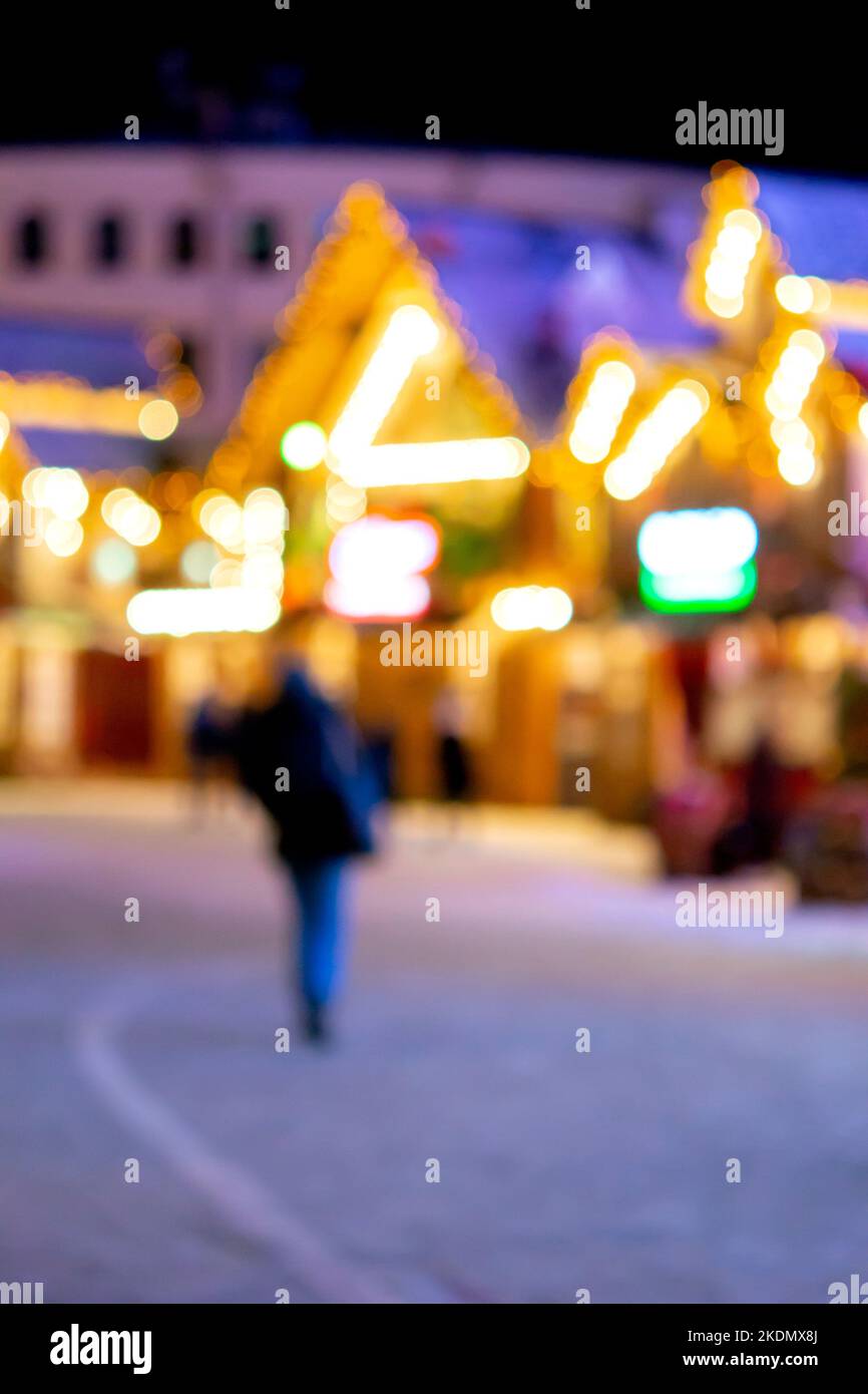 Sfondo sfocato. La gente cammina nella piazza della città la sera d'inverno. Silhouette nere di persone che camminano vicino a case decorate illuminazione luminosa. Luce bianca bokeh sfocare i punti dalle luci della casa incandescente Foto Stock