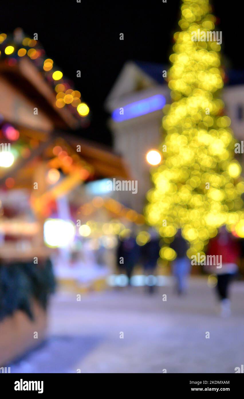 Sfondo sfocato. Albero di Natale di Capodanno decorato giallo ghirlande illuminazione, piccole case di legno ghirlande sul tetto e sagome di persone che camminano lungo la strada della città. Foto Stock