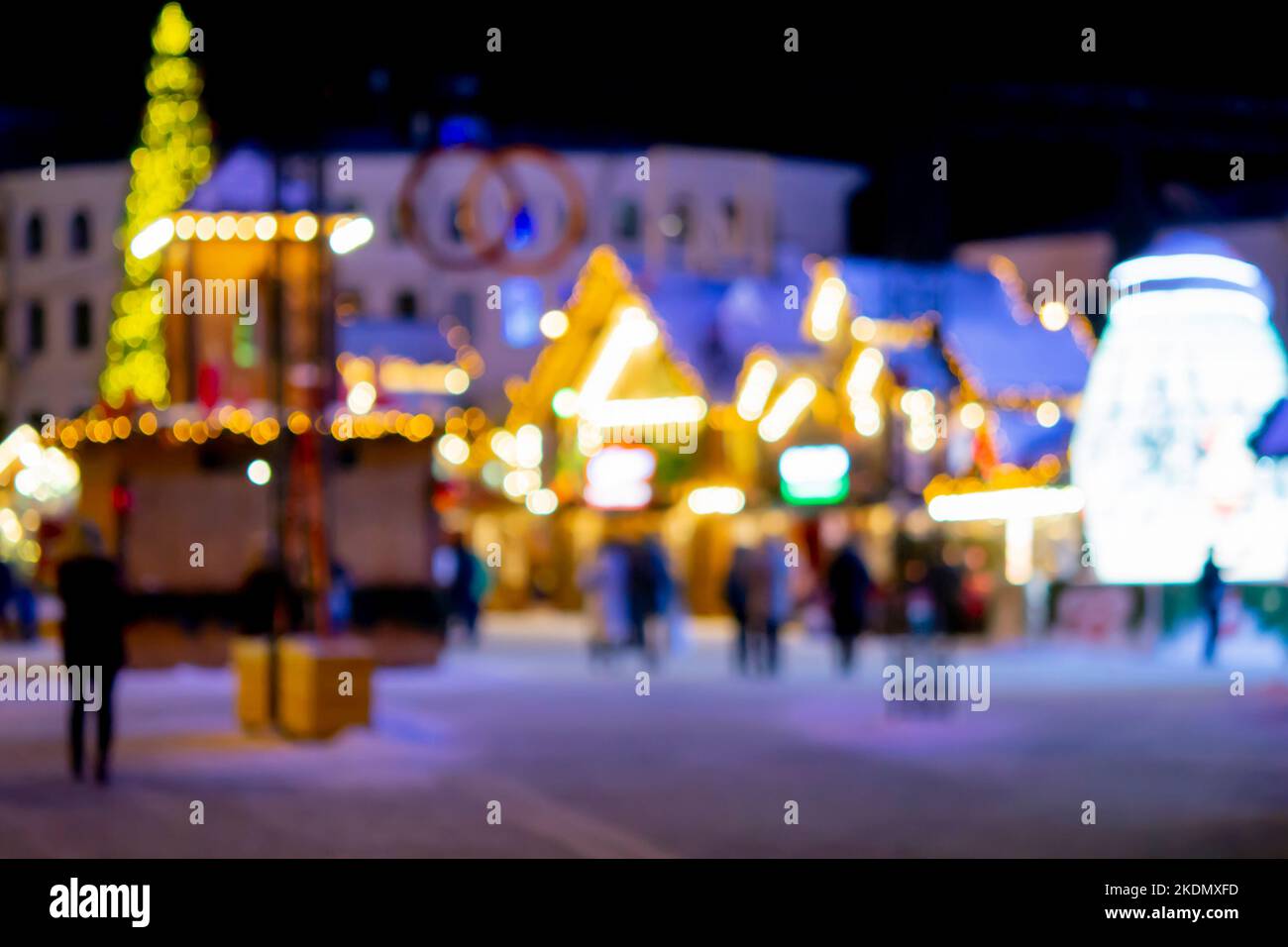Sfondo sfocato. La gente cammina nella piazza della città la sera d'inverno. Silhouette nere di persone che camminano vicino a case decorate illuminazione luminosa. Luce bianca bokeh sfocare i punti dalle luci della casa incandescente Foto Stock