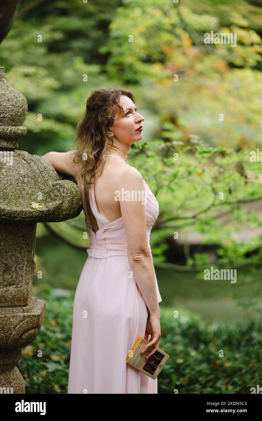 Donna che indossa un abito rosa che legge un libro in un giardino idilliaco Foto Stock