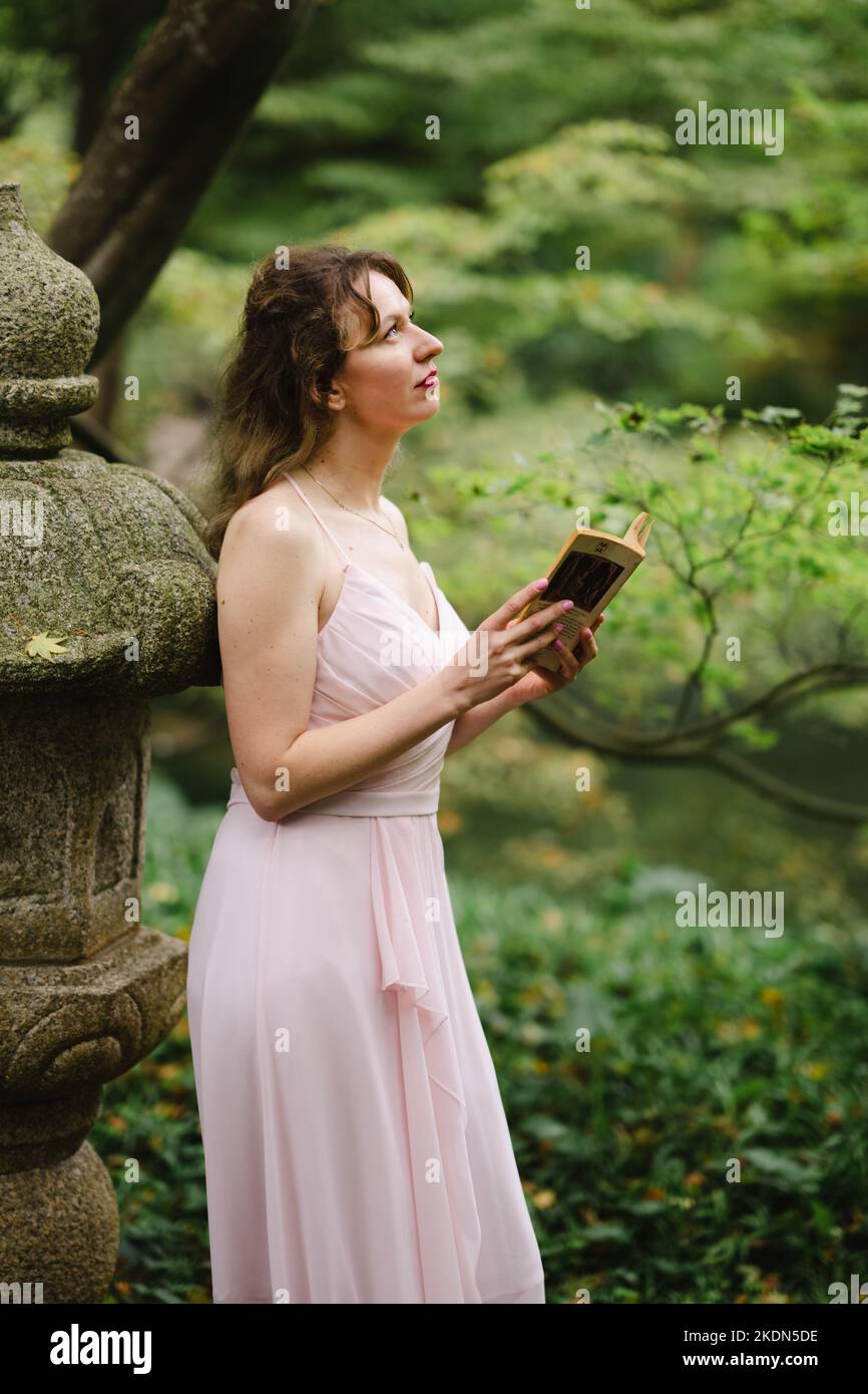 Donna che indossa un abito rosa che legge un libro in un giardino idilliaco Foto Stock