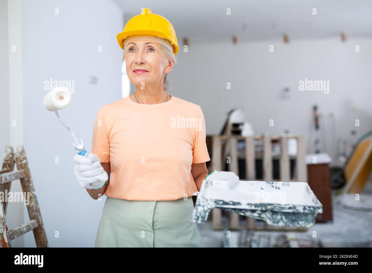 Ritratto di una donna anziana pittore in casco protettivo che dipinge le pareti con rullo di vernice Foto Stock