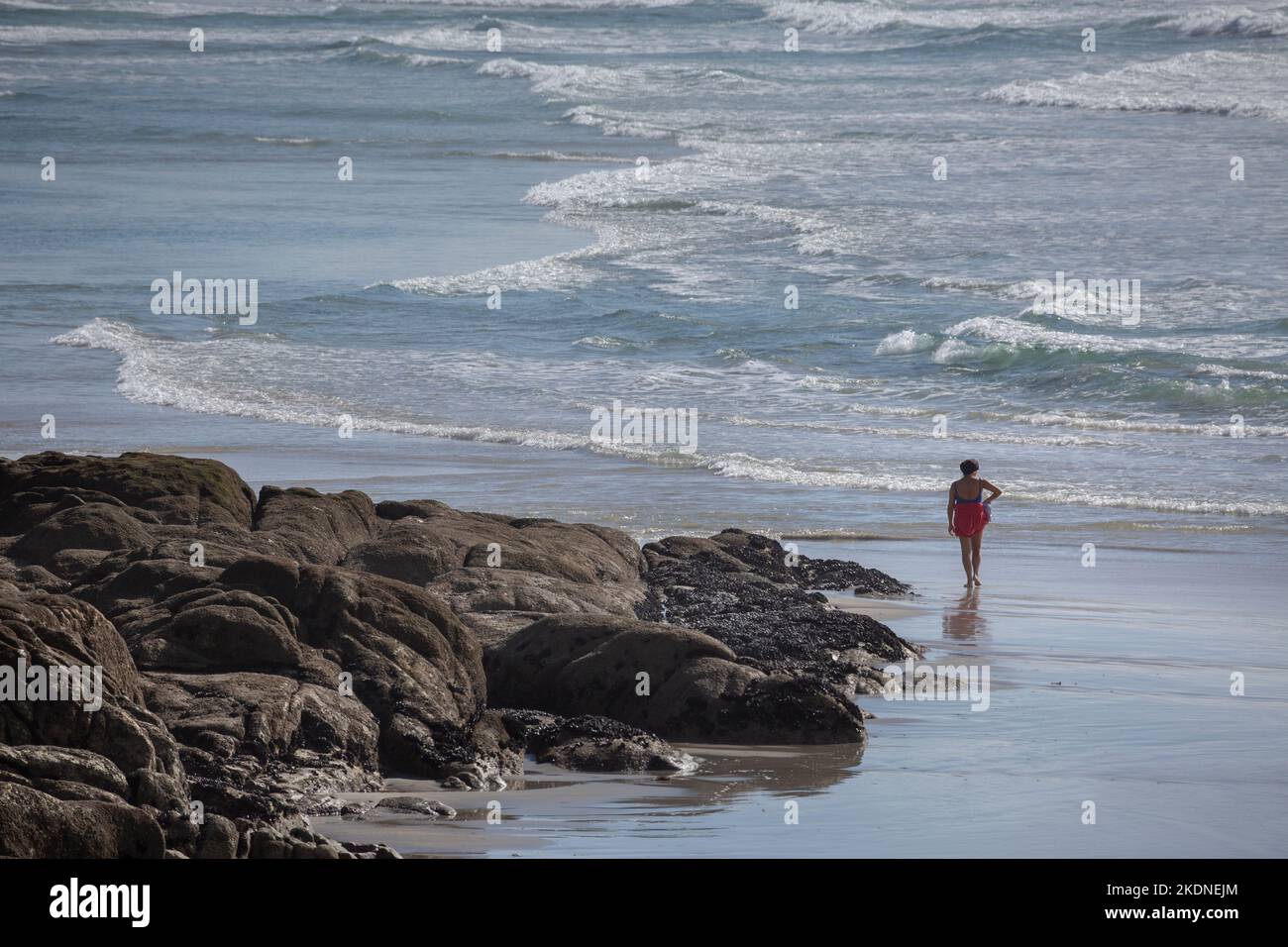 Una donna in costume da bagno nero e un sarong rosso cammina lungo la Playa de A Lanzada accanto alle rocce. O Grove, Galizia Foto Stock