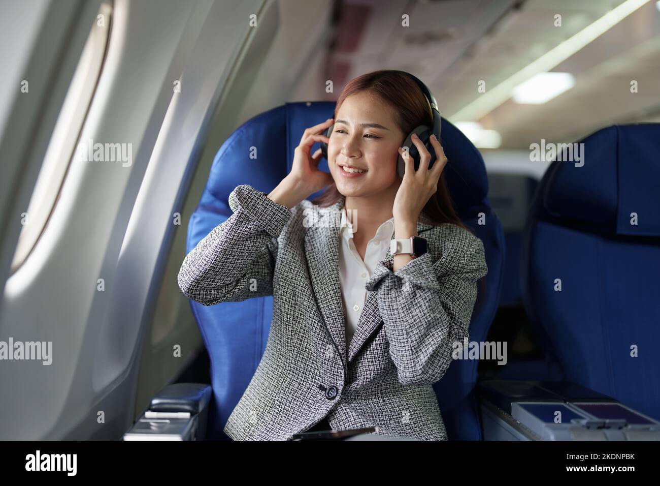 Ritratto di Una donna di successo asiatico business in un aereo si siede in un posto business class e utilizza un intorno-orecchie cuffie per la riproduzione di musica durante Foto Stock