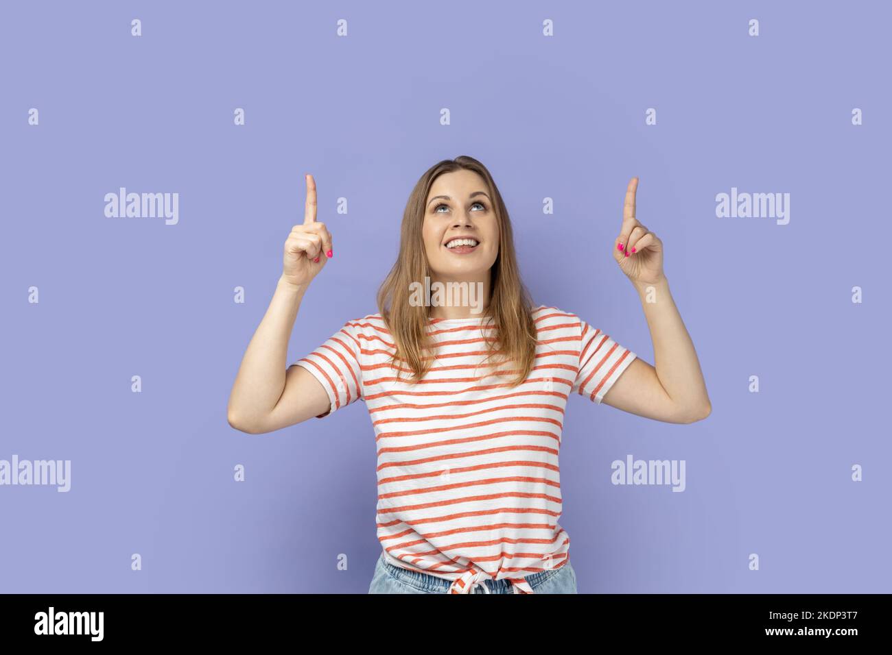 Guardate là, area pubblicitaria. Ritratto di una donna bionda sorridente che indossa una T-shirt a righe che guarda stupita e che indica uno spazio vuoto per la promozione. Studio al coperto isolato su sfondo viola. Foto Stock