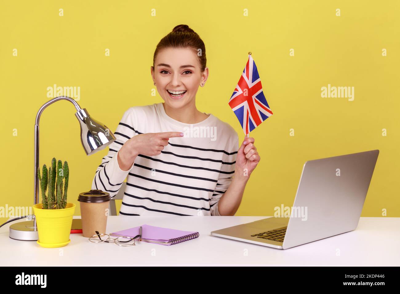 Sorridente donna positiva seduta sul posto di lavoro con, indicando la bandiera del Regno Unito di Gran Bretagna, esprimendo felicità, delocalizzazione all'estero. Studio interno girato isolato su sfondo giallo. Foto Stock