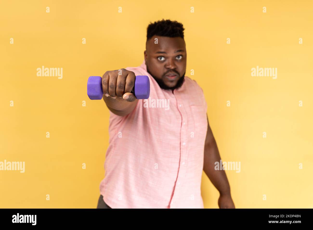 Ritratto di serio uomo portatore sicuro di sé indossando una camicia rosa allenando le braccia con il manubro, guardando la macchina fotografica, stile di vita sano. Studio al coperto isolato su sfondo giallo. Foto Stock
