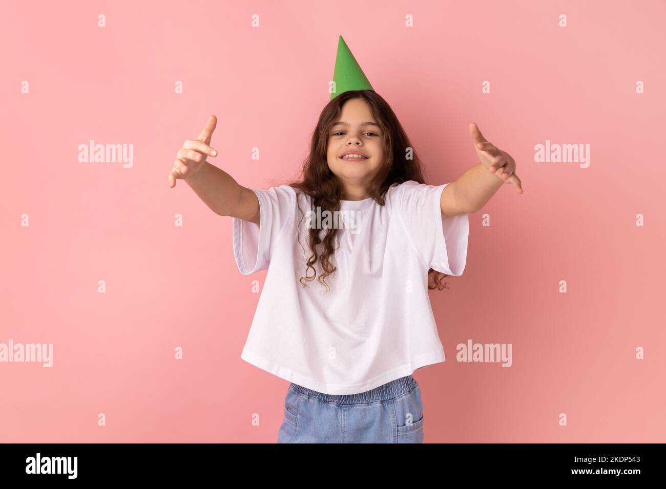 Ritratto di felicissima bambina che indossa una T-shirt bianca e un cono da festa che offre abbracci liberi con mani tirate, invitando ad abbracciare. Studio in interni isolato su sfondo rosa. Foto Stock