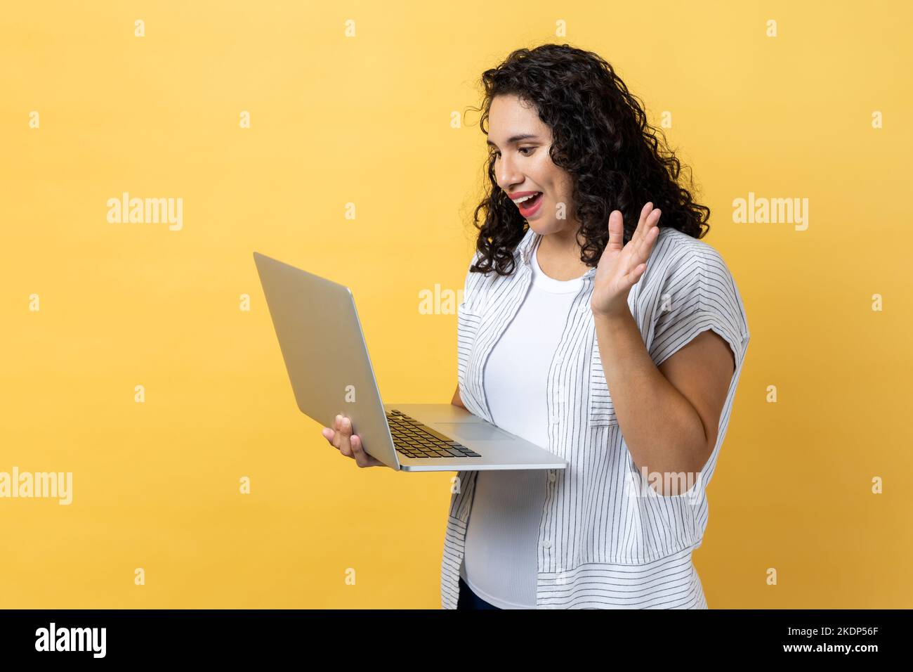 Ritratto di amichevole felice donna compiaciuta con capelli scuri ondeggianti lavorando sul portatile, videochiamare, agitare la mano, dire ciao o addio. Studio al coperto isolato su sfondo giallo. Foto Stock