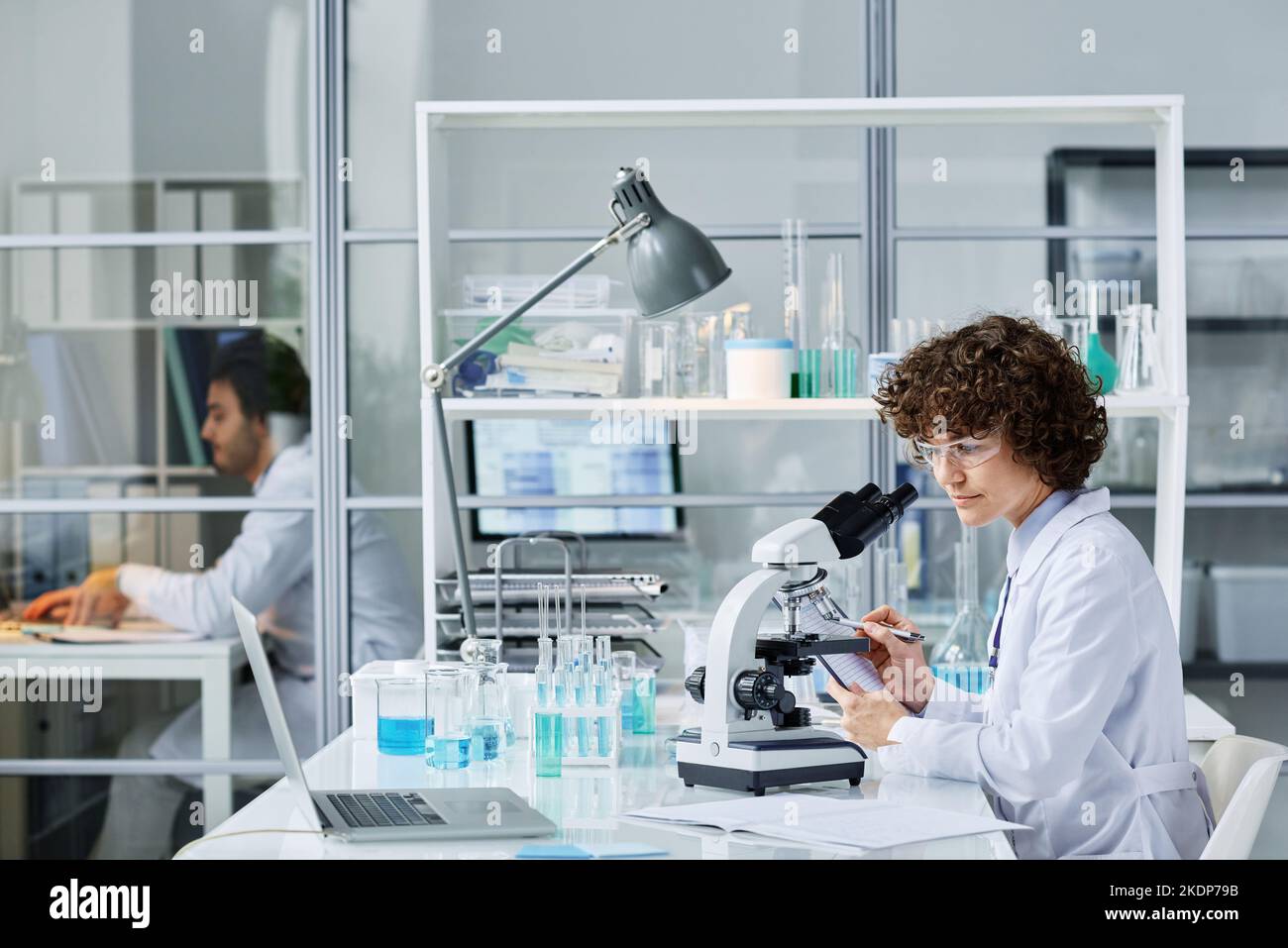 Giovane chimico o microbiologo in camice da laboratorio e occhiali protettivi seduti sul posto di lavoro con microscopio e prendere appunti durante la sperimentazione Foto Stock