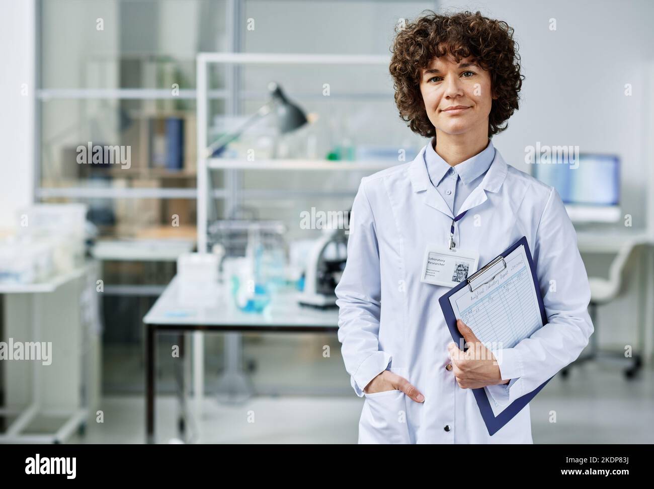 Medico giovane e sicuro con documento negli appunti davanti alla fotocamera in laboratorio scientifico contro il suo posto di lavoro Foto Stock