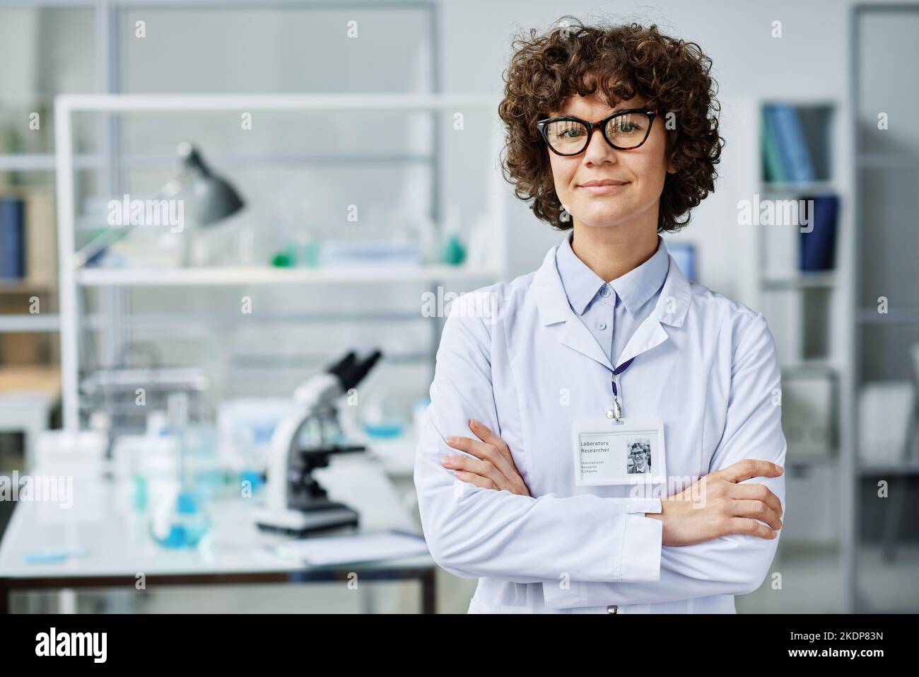 Giovane donna di successo brunette clinico o scientifico esperto in camice da laboratorio e occhiali incrociando le braccia al petto e guardando la macchina fotografica Foto Stock