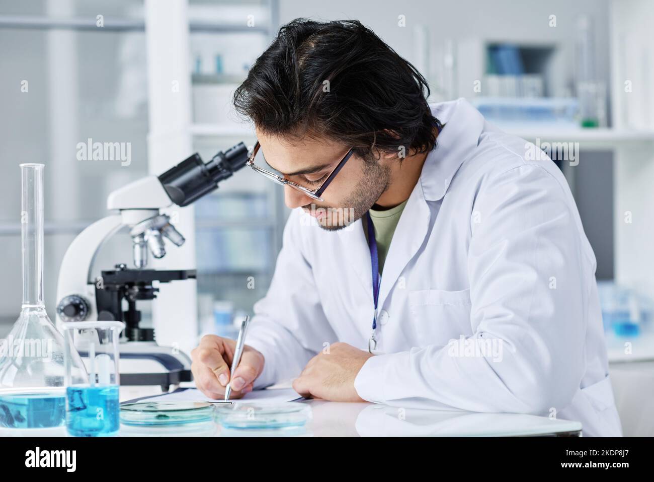 Giovane scienziato moderno in camice da laboratorio e occhiali che prende appunti nel documento mentre scrive i risultati dell'esperimento clinico Foto Stock
