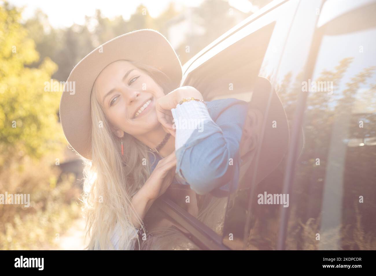Gioioso sorridente premurosa donna bionda in cappello e jeans vestito appoggiato fuori dalla finestra di auto gioia viaggio estate natura alla luce del sole Foto Stock