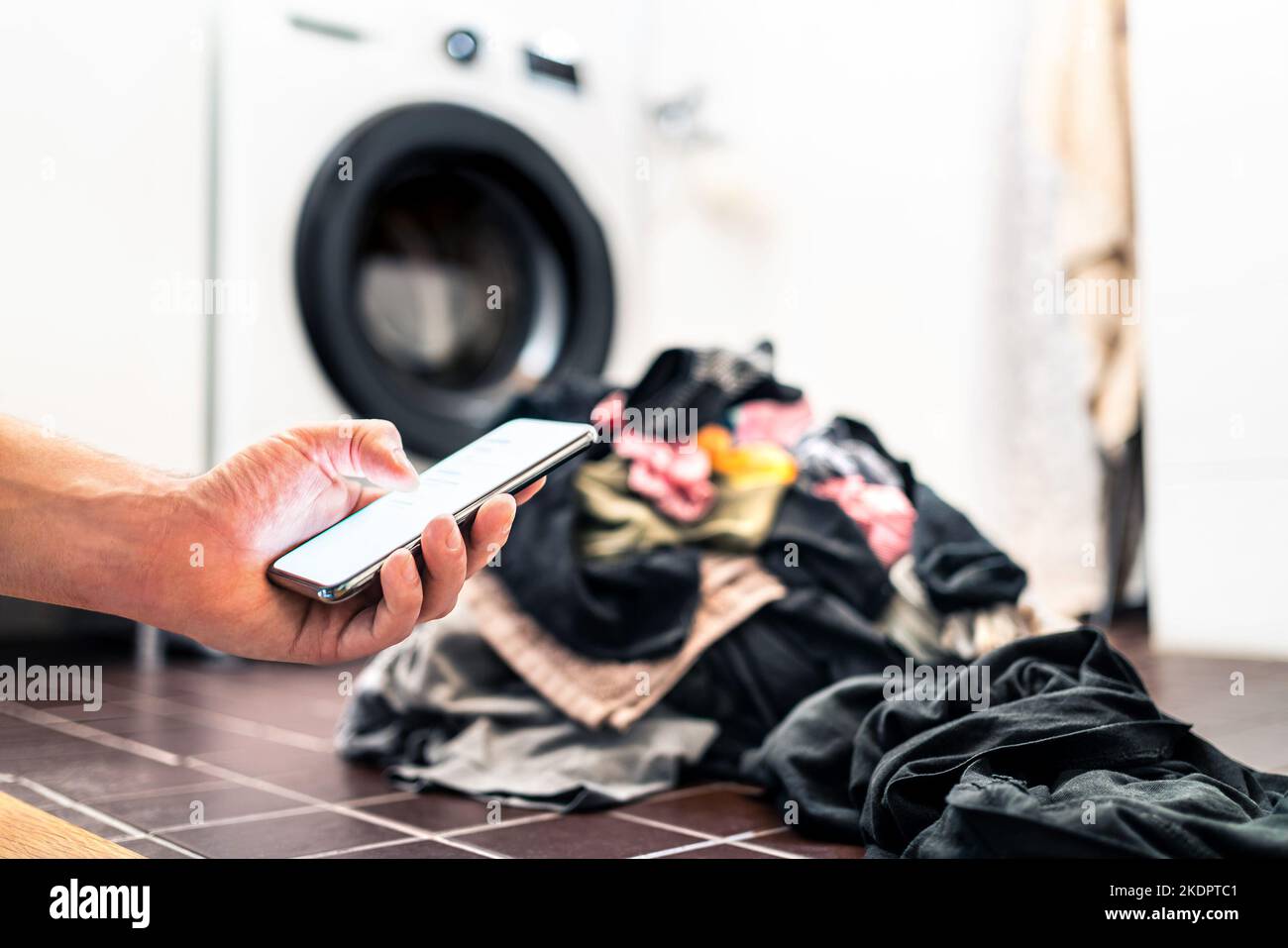 Lavatrice, telefono e lavanderia. Tecnologia per la casa intelligente e smartphone. Persona che usa il cellulare o SMS mentre fa i lavori della casa. Foto Stock