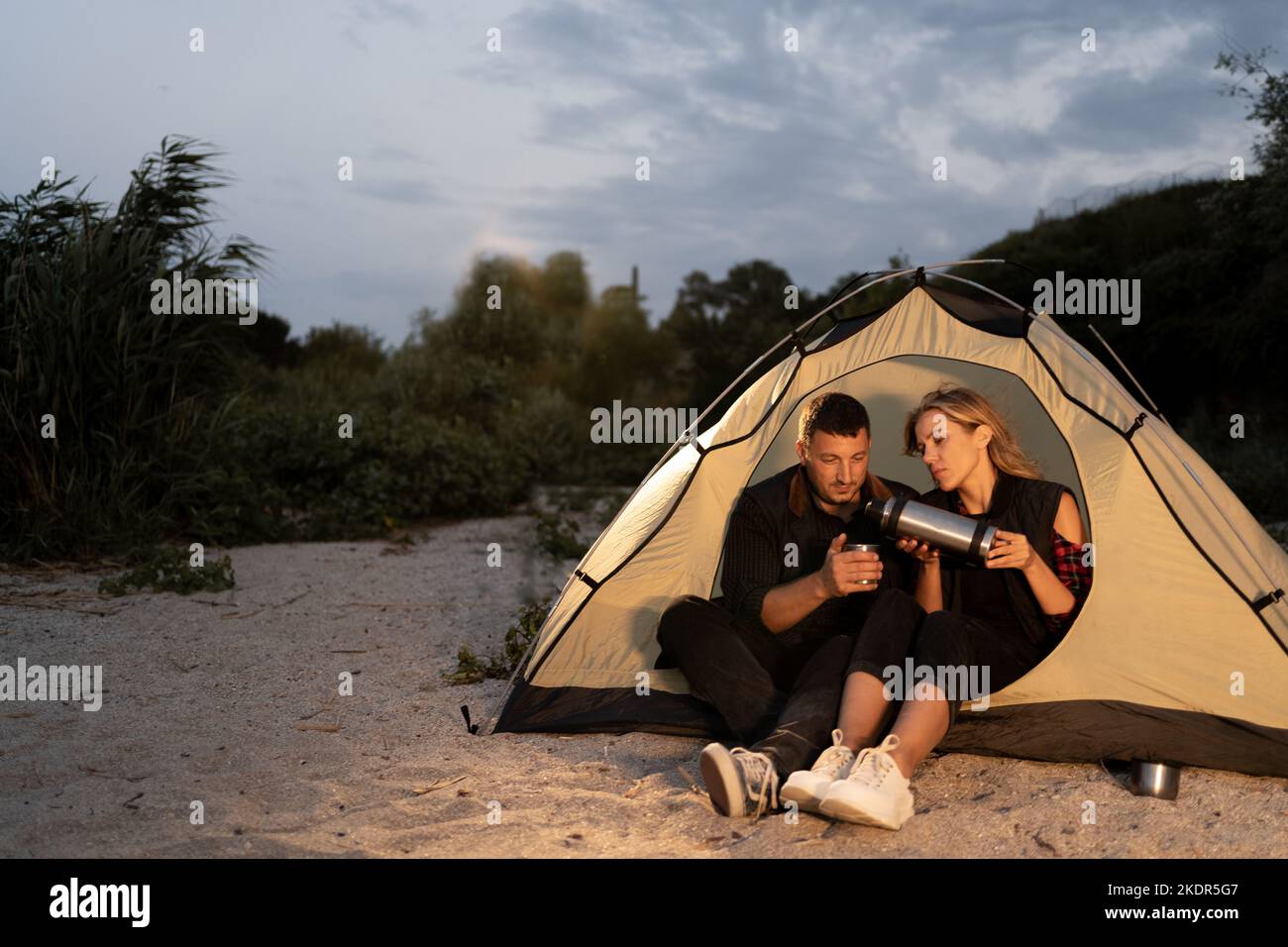 Campeggio notturno sulla spiaggia. I turisti uomini e donne riposano di fronte ad una tenda sotto il cielo nuvoloso della sera, bevendo caffè sulla spiaggia e. Foto Stock