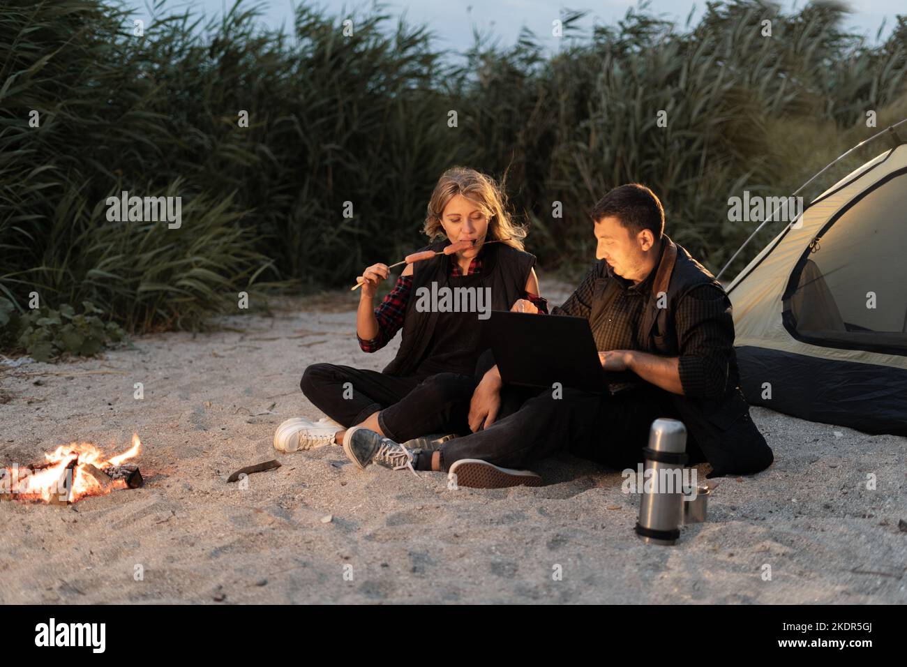 Una coppia che usa un computer all'esterno di una tenda per un campeggio in spiaggia la sera d'estate. Rilassarsi in tenda da campeggio vicino al mare al tramonto. Freelance Foto Stock