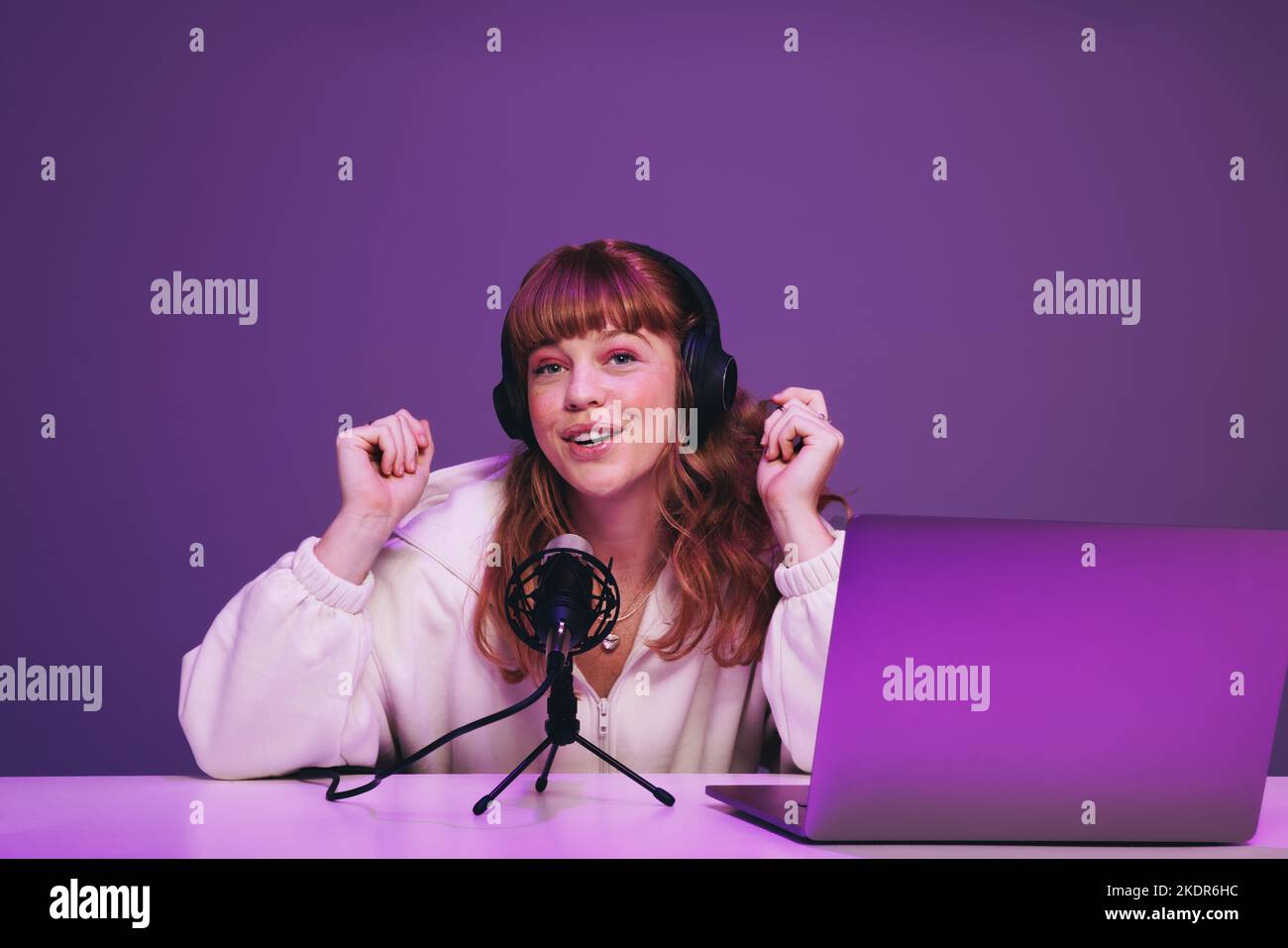Buon giovane podcaster che parla in un microfono in uno studio di registrazione. Giovane donna che ospita una trasmissione audio dal vivo su uno sfondo viola. Donna Foto Stock