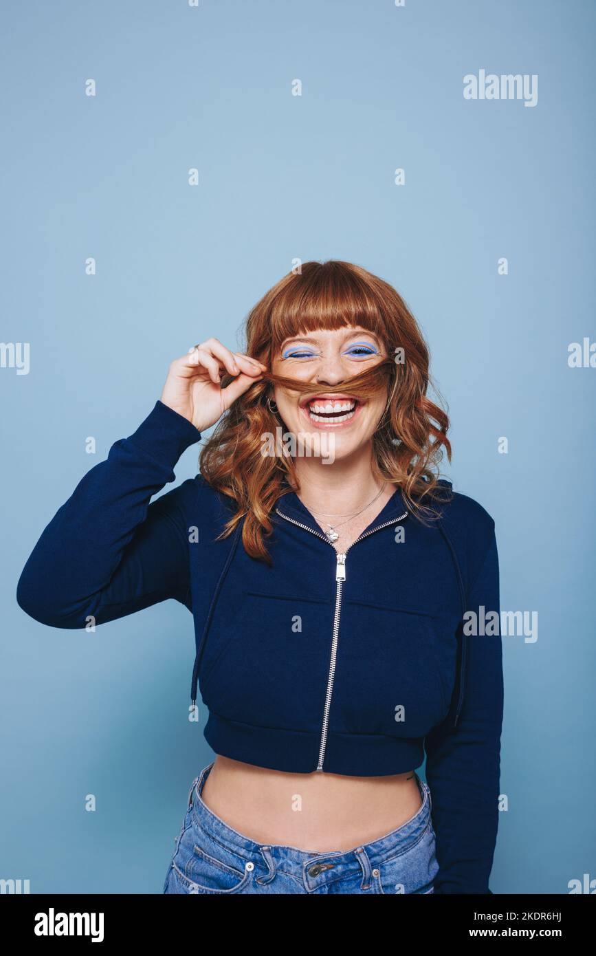 Giovane donna allegra che ride felicemente mentre fa un baffo con i suoi capelli in uno studio. Giovane donna allegra che si diverte in piedi contro un blu Foto Stock