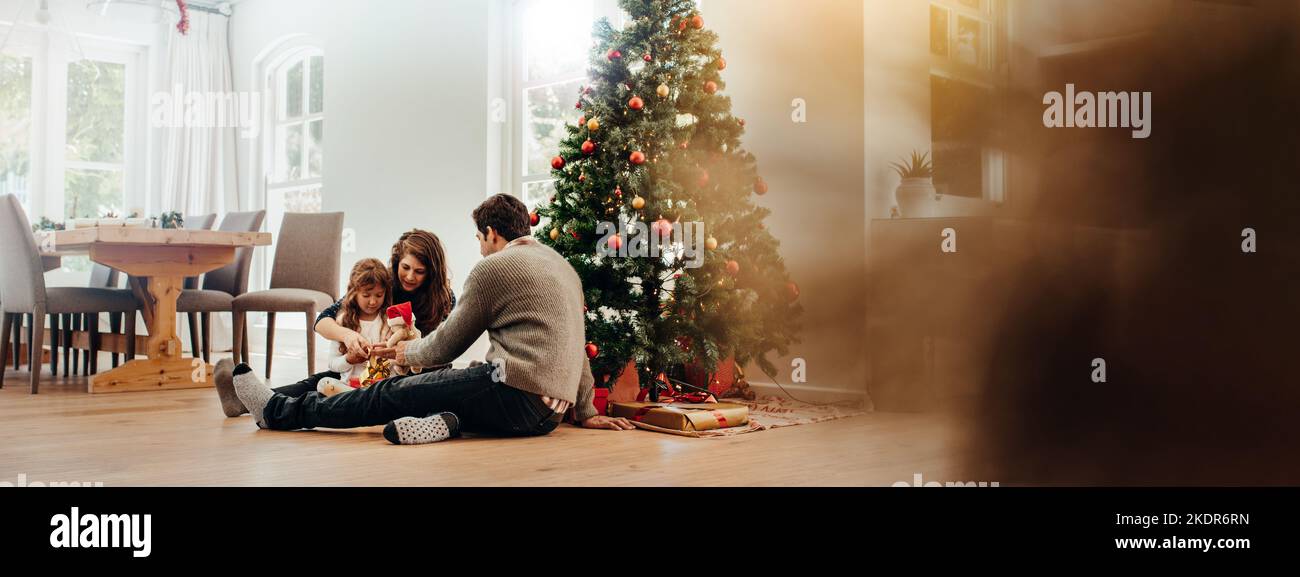 Famiglia seduta vicino ad albero di Natale apertura di confezioni regalo. Coppia giovane aiutando la loro figlia aprire i regali di Natale. Foto Stock