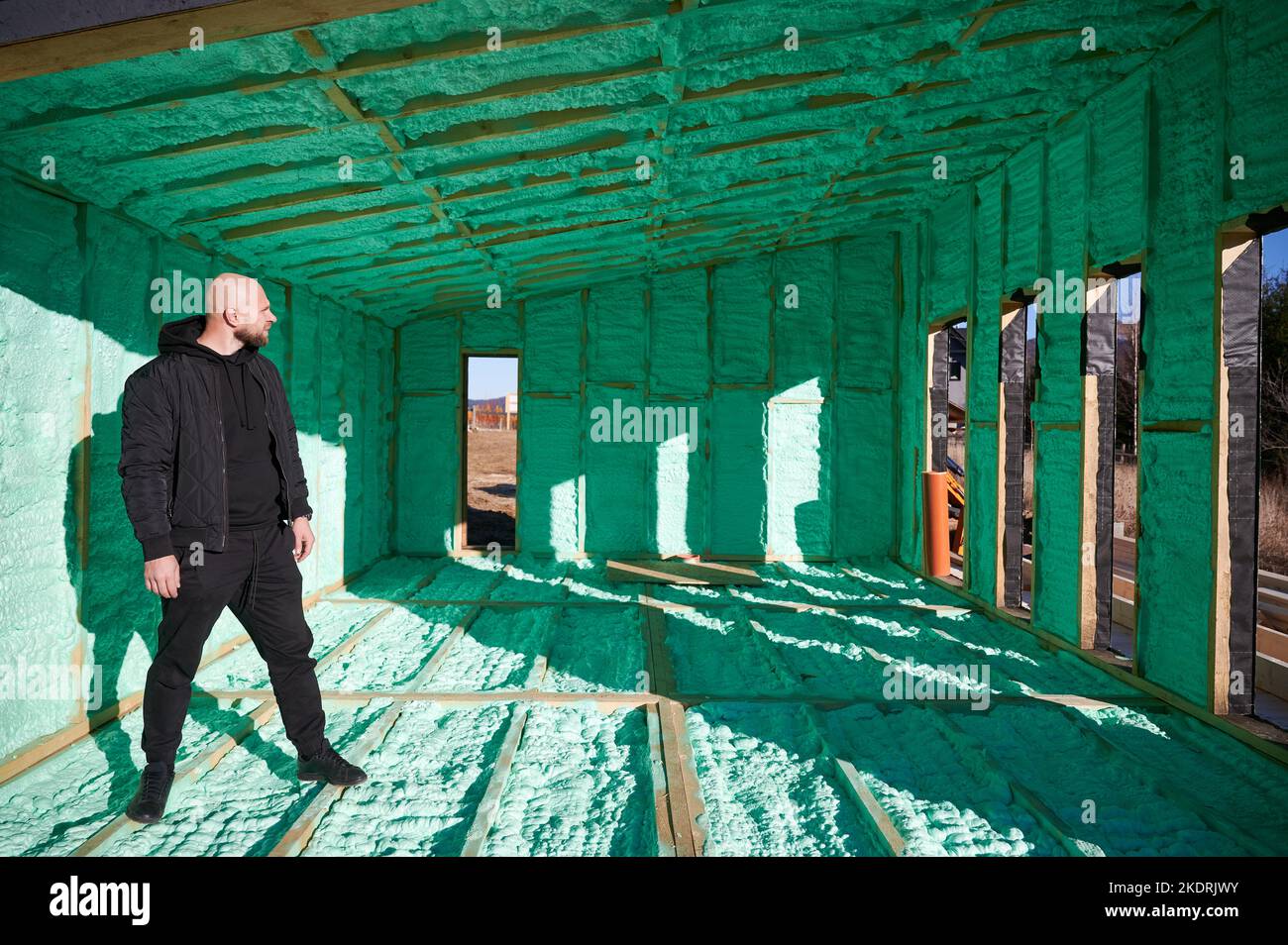 Sviluppatore maschio edificio casa con struttura in legno. Uomo calvo in piedi in cantiere, ispezionando la qualità dei lavori di isolamento con schiuma poliuretanica sulle pareti. Foto Stock