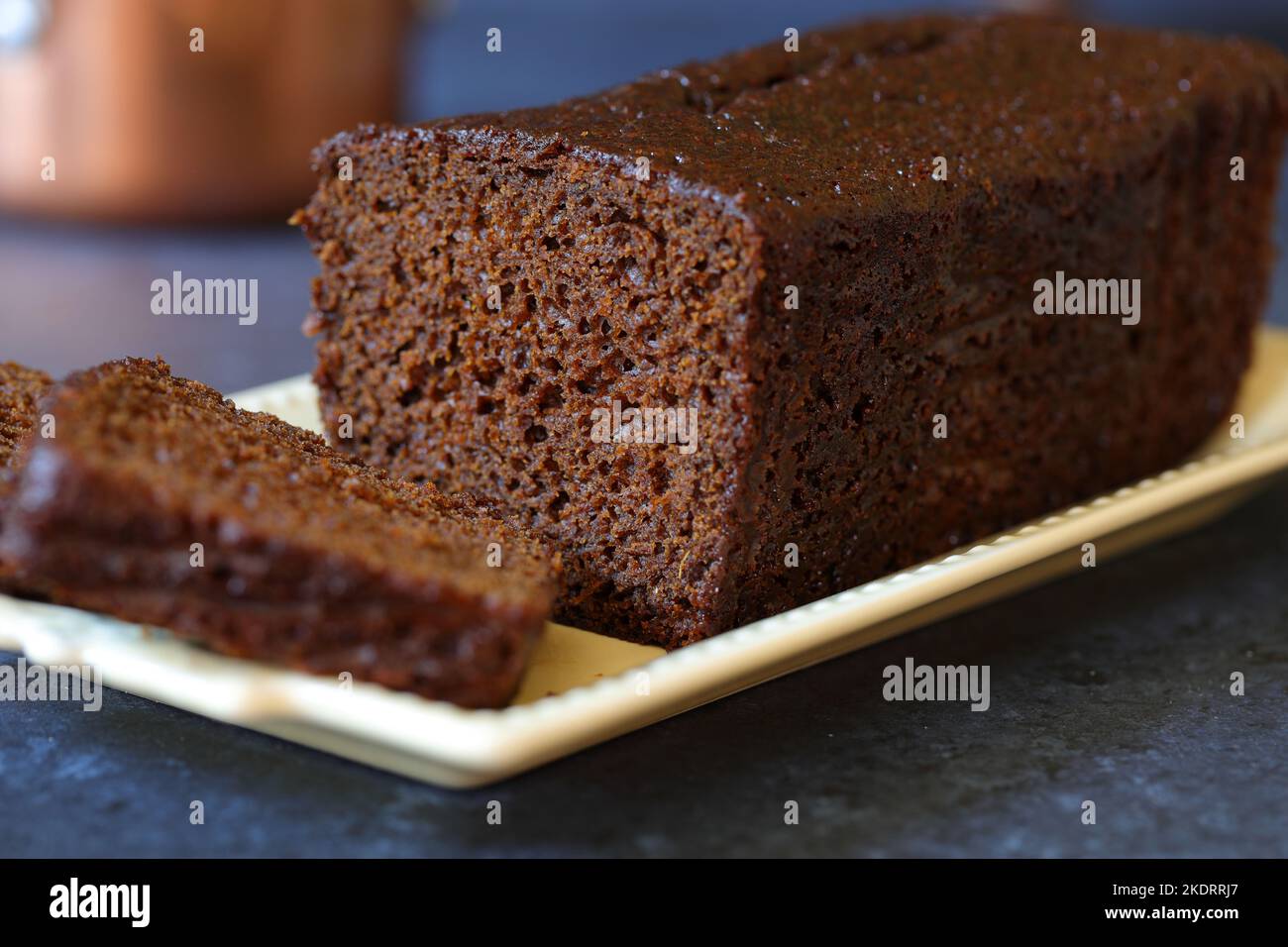 Torta di zenzero giamaicano affettato in casa scuro e piccante zenzero e torta di focaccia speziata su un piatto di servizio oblungo crema d'epoca decorativo sfondo blu scuro Foto Stock
