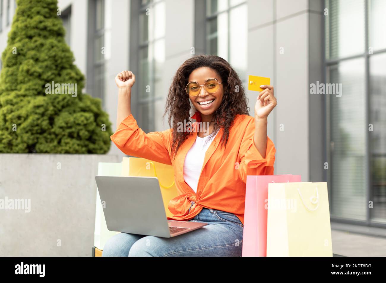 Soddisfatto giovane donna nera con carta di credito, borse e pc godere di shopping online, alza le mani, gioisce in offerta Foto Stock