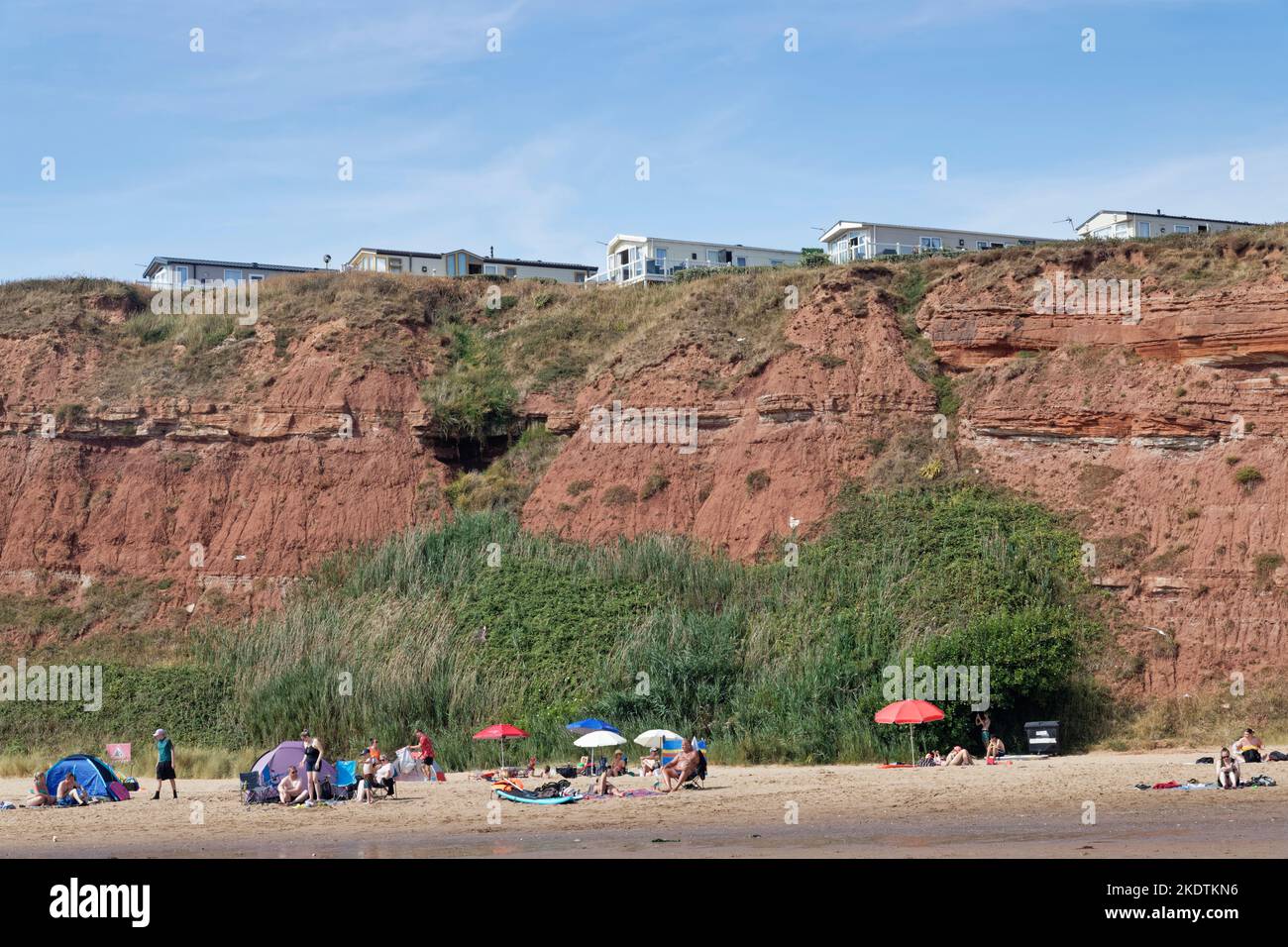 Turisti a Sandy Bay Beach con caravan statico parco vacanze arroccato sulle cime rosse della scogliera di arenaria sopra, Exmouth, Devon, Regno Unito, agosto. Foto Stock